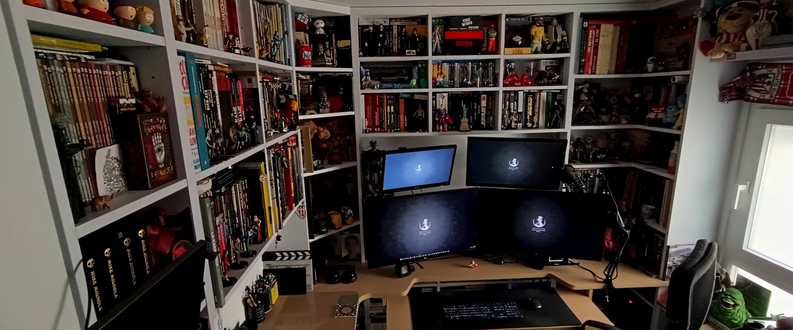 Escritorio con tres monitores en un rincón de estudio rodeado de estanterías con libros y figuras
