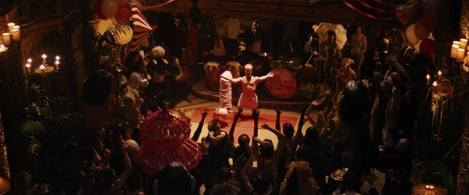 Fiesta nocturna en el interior de la mansión de Babylon, con bailarines y percusionistas alrededor de un hombre semidesnudo en el centro de la pista.