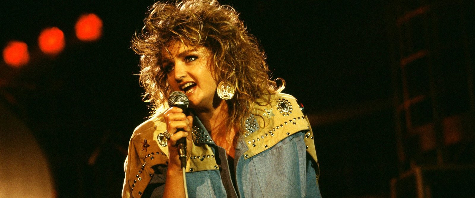 Bonnie Tyler con media melena rizada y cazadora vaquera con tachuelas, cantando en directo en los años ochenta