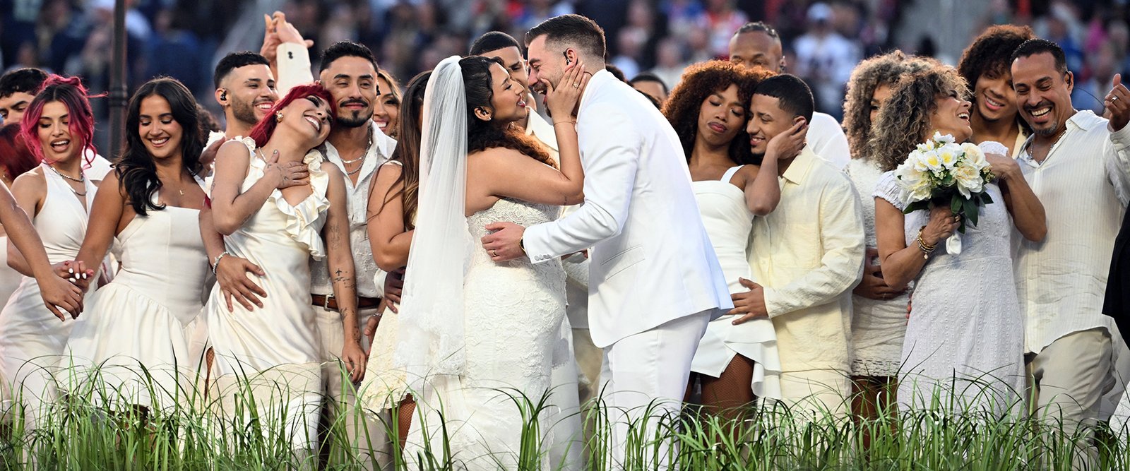 Una pareja de novios se besa rodeada de invitados vestidos de blanco y con hierba de caña de azúcar en primer plano, durante la boda del espectáculo de Bad Bunny