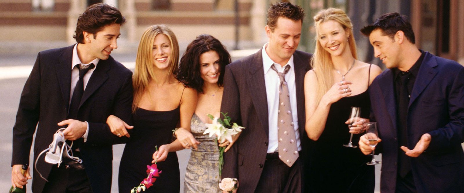 El reparto de Friends caminando sonriente por la calle en una escena de la serie, con Matthew Perry en el centro, de traje y corbata