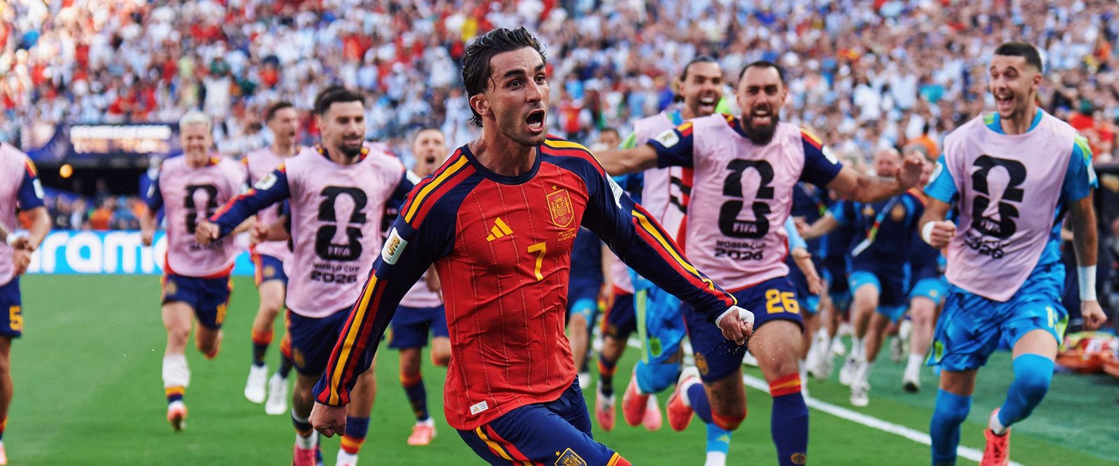 Un jugador de España celebra corriendo con el puño en alto, compañeros de fondo con petos rosas de calentamiento con el logo oficial del Mundial 2026