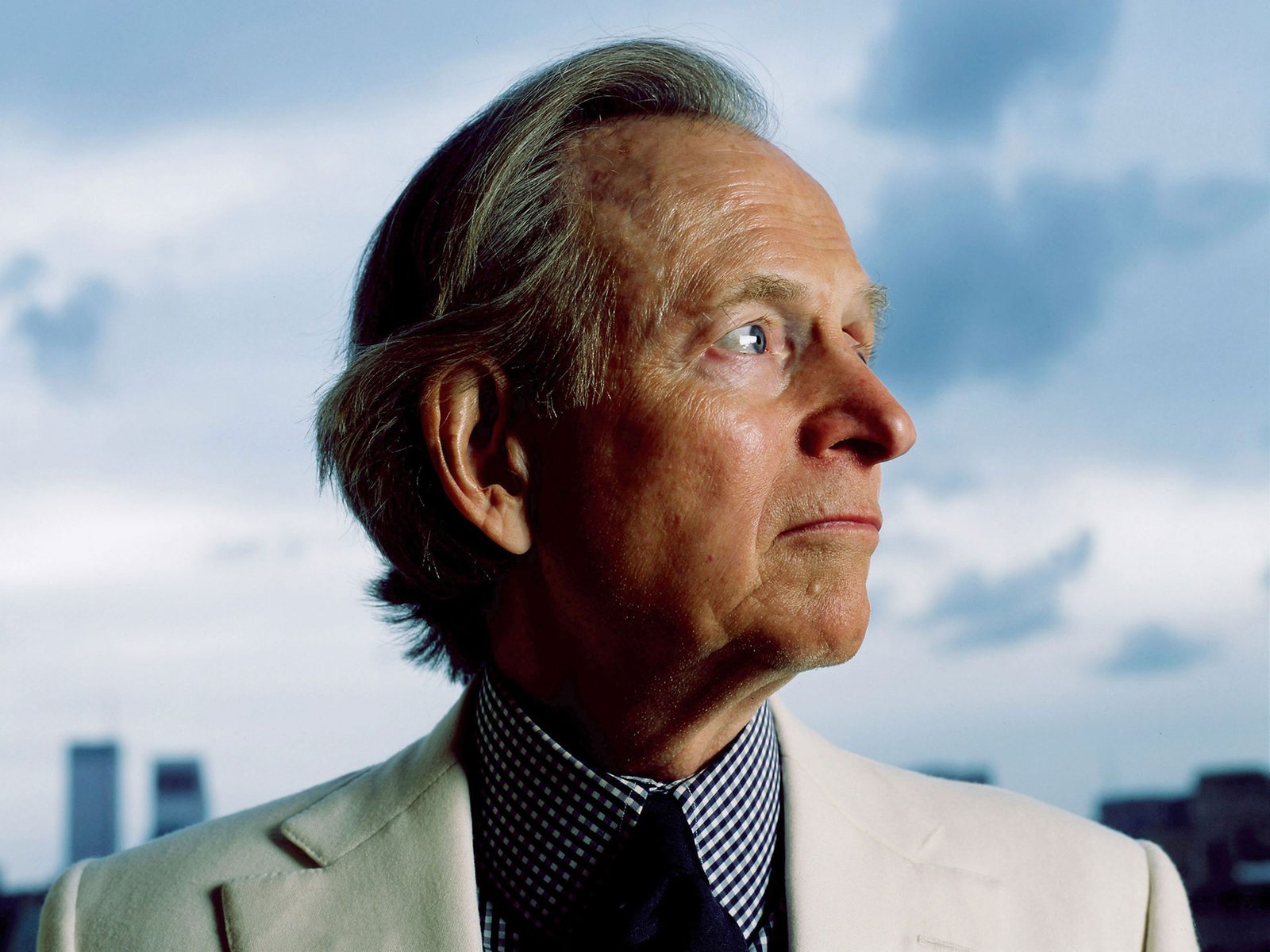 Tom Wolfe en perfil, con traje claro, mirando al horizonte de Nueva York bajo un cielo nuboso