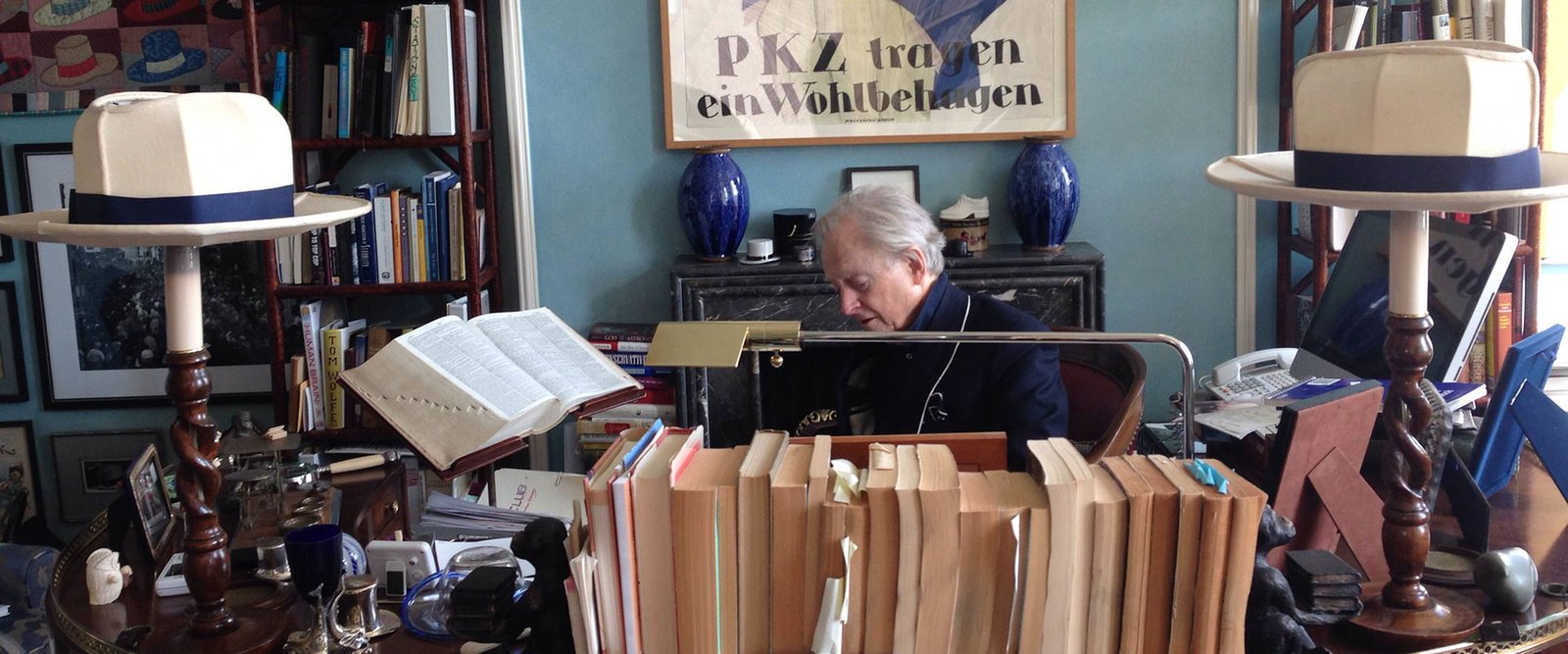 Tom Wolfe sentado a su escritorio abarrotado de libros, entre dos sombreros claros y un cartel antiguo en alemán