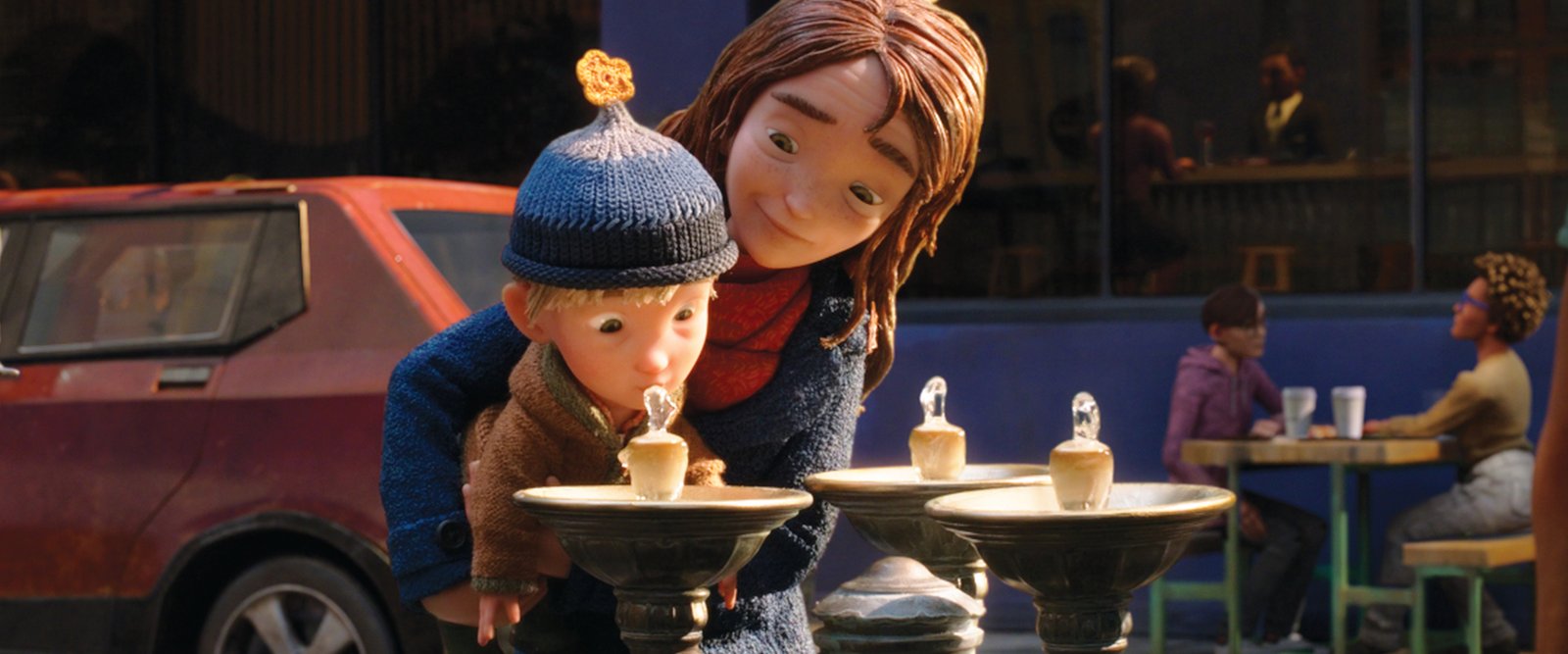 Una niña de pelo castaño sostiene a un niño pequeño con gorro de lana para que beba de una fuente pública, junto a un coche rojo y una terraza.